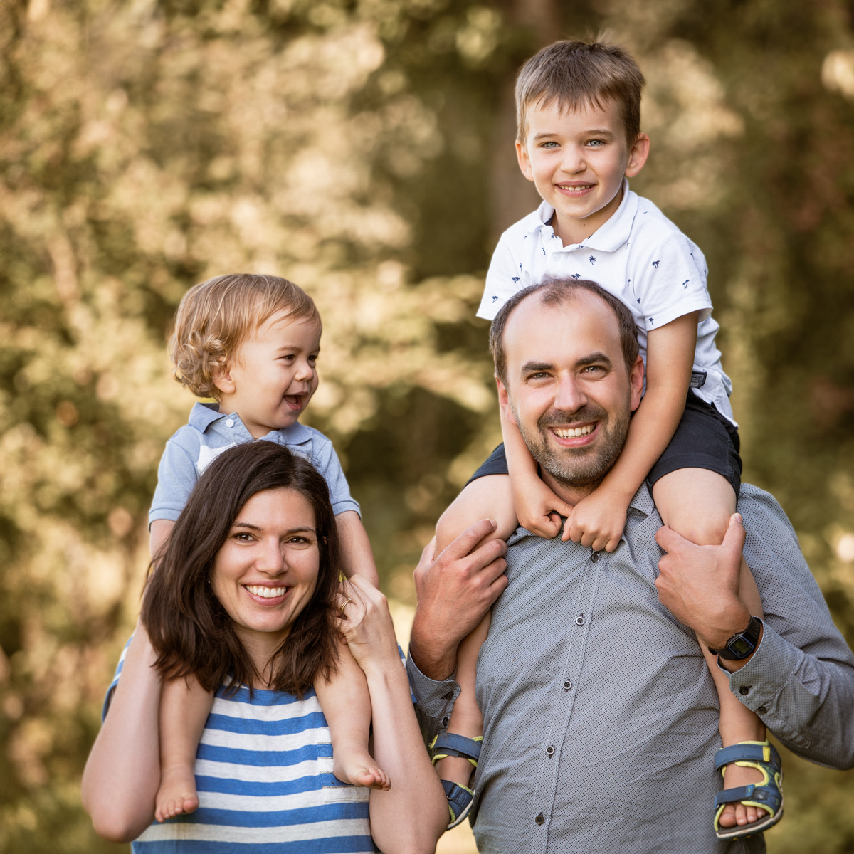 Lachende Familie im Freien; beide Eltern tragen jeweils ein Kind auf den Schultern, alle wirken gelöst und fröhlich.